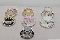 Full view of seven porcelain floral teacup and saucer sets arranged on a white surface showing different colors and patterns.