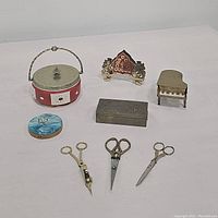 Overview of all items in the lot arranged on a white surface to show scale and detail including the jar, powder compact, hinged box, miniature piano, crest plaque and scissors.