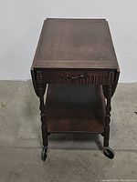 Front view of vintage wooden drop leaf serving table with drawer and lower shelf, showing carved details and caster wheels.