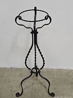 Front view of the wrought iron plant stand displaying twisted central column, ring holder and three footed legs on concrete floor.