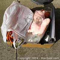 View of assorted items arranged in a cardboard box including the plush toy, photo album, curtain rings, glasses, corded trouble light wrapped in plastic, and others.
