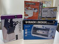 Photo of boxed kitchen items: Oneida 16-piece knife set with block, Proctor Silex slow cooker, Black & Decker toaster oven, Premium hand mixer.