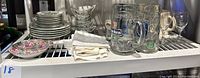 Full lot showing stacked J.P.L France plates and bowls, napkins, clear glassware, and beer mugs grouped on a shelf
