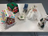 Table setup showing the ceramic gingerbread house with colorful candy details, green knitted Santa tissue box holder, blue wooden sleigh, Santa head decoration, snowman soap dispenser, and white tree dish.