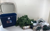 Blue storage bin open with artificial green Christmas garland, string lights, timers, extension cords, and mesh ribbon on a flat surface next to it.