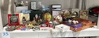Wide view of all assorted Christmas ornaments including boxed collectibles, metal, ceramic, and glass pieces arranged on a white surface under sunlight.