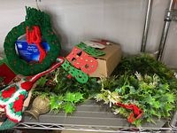 Shelving unit with green plastic wreath adorned with red berries, cardboard box containing string lights, and greenery wreath with pinecones and red ribbon.