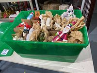 Green plastic tote filled with assorted Christmas ornaments, many wrapped in brown tissue paper, containing Santa and snowman figurines, and ice skate ornaments.