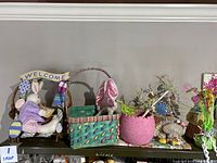 Photo of assorted Easter decorations on a shelf including plush bunny in woven basket with 'Welcome' sign, green basket with bunny ear, pink planter with flowers, and other floral and bunny decorations.