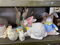 Shelf displaying various ceramic Easter bunny figurines and related decorations including large and small bunnies in pastel colors and some small egg and chick shaped pieces.