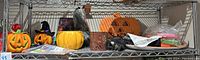 Shelf with Halloween decorations including plastic and light-up pumpkins, ceramic and metal candle holders, black fabric cape, pumpkin stickers and plush spider.