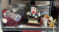 Holiday basket with assorted holiday decor including plush bear, snowmen figurines, bagged pine cones, sleigh, pillow and stocking hanger shown on shelf
