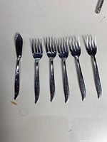 Six silver forks and one silver spreader knife, showing signs of tarnishing, arranged on a light surface.