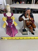 Two porcelain figurines, a woman in a purple gown with yellow bows and a seated man reading beside stacked books.