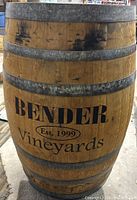 Front view of wooden wine barrel showing 'Bender Vineyards Est. 1999' text and metal bands, with some scratches and dark marks.