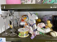 Wooden painted Easter figures including bunny, chick, lamb, along with clear glass candle holders, spool of Easter ribbon, and butterfly decoration on green shelf.