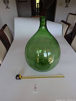 Front view of a large green glass demijohn placed on a white surface with a measuring tape laid out showing scale of approx 18 inches diameter and 25 inches tall.