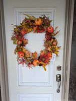 Photo showing full 26 inch autumn wreath hanging on a white front door, decorated with orange pumpkins, red and yellow artificial leaves, pine cones, and flowers.