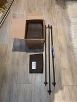 Dark brown blackout curtain panels folded and packed, with three black adjustable curtain rods placed next to them on the floor.