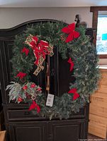 Front view of the Christmas wreath hanging on a dark armoire, showing red bows, mesh ribbon, and frosted leaf and berry accents.
