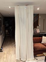 Long cream colored window panel hanging with visible tag, shown in living room setting on curtain rod.