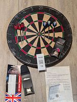 Full view of the dart board mounted on floor with two sets of darts and cases.