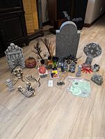 Multiple Halloween decorations arranged on floor including large tombstones, skull, pumpkin, skeleton hands, spiders, and other spooky props.