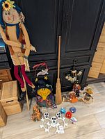 Assortment of Halloween decorations including scarecrow, witches broom, witch figure, small scarecrow, multiple ghost and pumpkin figurines arranged on floor near black cabinet.