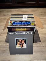 Photo of roughly 40 assorted LP vinyl records in a cardboard box, visible titles include ABBA Greatest Hits Vol. 2 in front and a Blackbuster disco album inside the box.
