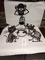 Three metal candle holders shaped as Halloween skeleton characters: Mr. Bonesy standing with hands on skull head, cat figure, and dog figure linked by chain. All black metal, designed for votive candles.