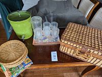 Photo showing green beverage dispenser with spigot, clear plastic glasses and pitchers, wicker plate holders, and wicker basket closed with leather strap on wood table.