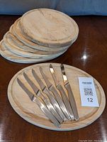 Six oval maple steak boards stacked with one displayed next to six J.A. Henckels steak knives on a wooden surface