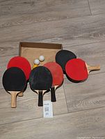 Six Eastpoint table tennis paddles showing red and black rubber surfaces and wooden handles, arranged on a wood floor with a box behind them containing four balls (three white, one orange).