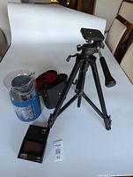 Items including black tripod, blue coin sorter, binoculars in red case, and leather wallet displayed on white backdrop