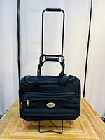Front view of the black American Tourister travel bag with extended trolley handle on wooden surface, showing front zip pocket and brand plaque