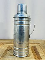 Full view front of the aluminum thermos showing shape, screw-top cap, and attached handle.