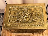 Top view of the rectangular box showing the embossed brass panel with a detailed scene of a family group in an interior setting with fireplace, furniture, and household objects.