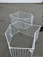 White metal baby gate assembled outdoors, showing multiple interlocking vertical bar panels forming an enclosed playpen shape.