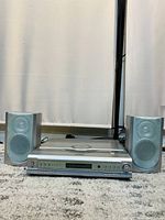 Front view of silver digital audio main unit with two matching speakers on carpeted floor in front of a curtain.