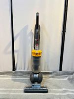 Front view of Dyson upright vacuum cleaner showing yellow cyclone and transparent dust container with handle and base with wheels.