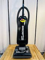 Front view of black Eureka Mega Boss upright vacuum cleaner on wooden surface with hose attached, showing model name and control panel on base.