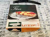 Photo of the Luminarc Fleur glass cake stand in its original packaging showing box design and brand label.