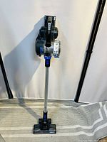 Full view of the vacuum cleaner standing upright showing the stick design with floor brush attachment, metal tube, and main vacuum body.