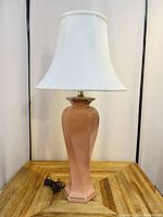 Front view of the vintage pink ceramic table lamp with white bell-shaped fabric lampshade and electrical cord placed on wood surface.