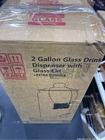 Cardboard box showing text 2 Gallon Glass Drink Dispenser with Glass Lid, extra durable, with a simple line drawing of the glass dispenser with lid and spigot.