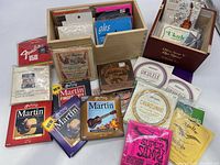 Wide shot showing multiple unopened packs of guitar and ukulele strings arranged on a wooden surface and inside small boxes, highlighting brands such as Martin, Dean Markley, Fender, Ernie Ball, D'Addario.