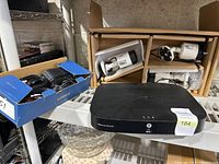 Wide photo showing the Lorex DVR on a shelf with a box containing four cords and three security cameras in cardboard packaging above it.