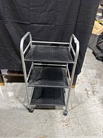 Front view of the metal utility cart showing three black perforated metal shelves and tubular silver metal frame.