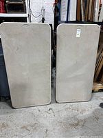 Two Samsonite folding tables standing upright showing their beige plastic tops with rounded corners and carrying handles molded into the surface.