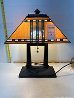 Frontal view of the lamp showing the stained glass shade with colorful geometric pattern and pull chain, with a measuring tape indicating 16 inches width.
